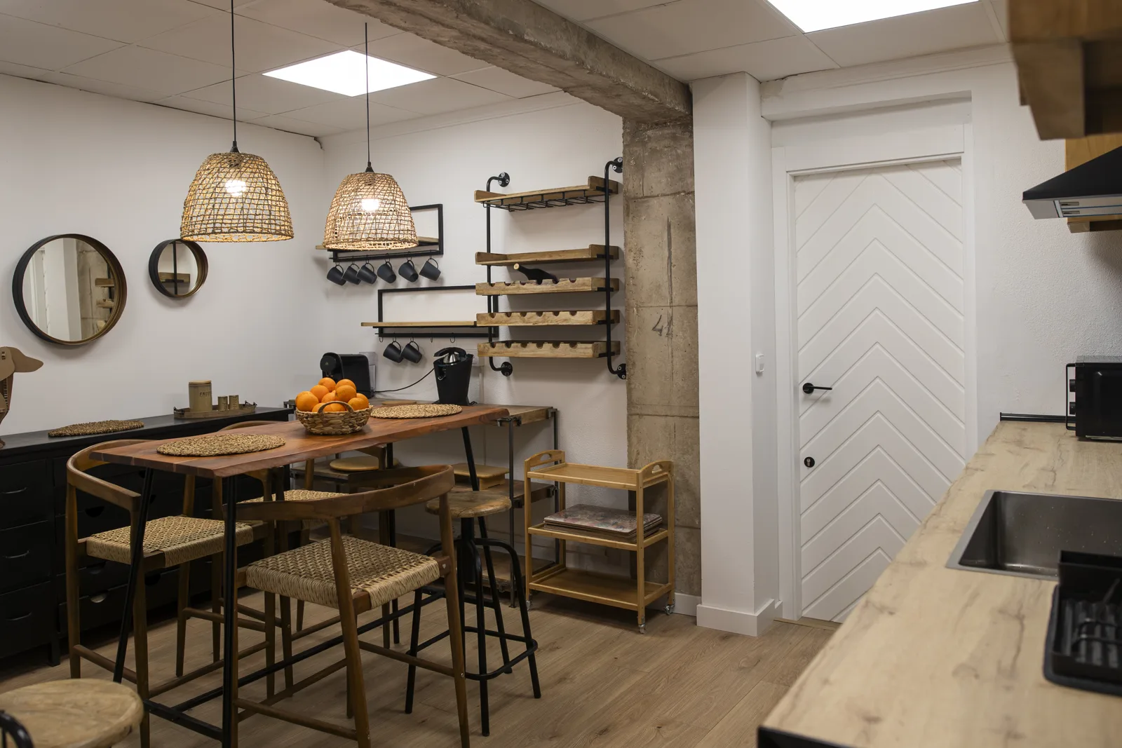 Cocina del Centro de Día con barra alta, taburetes de madera, lámparas de mimbre y estantes con menaje