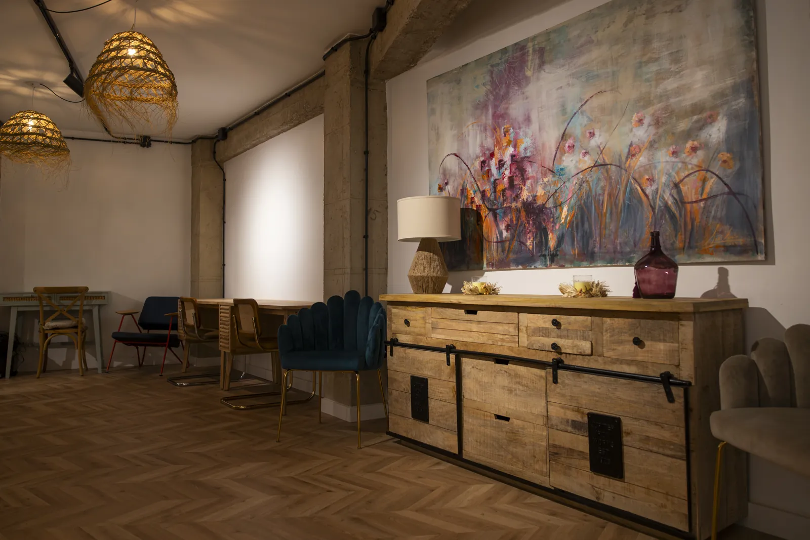 Sala del Centro de Día con suelo de madera, lámparas de ratán, un gran cuadro de flores sobre un aparador y sillas de terciopelo