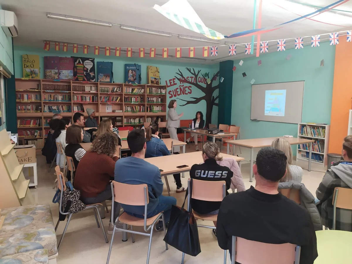 Charla para familias en la biblioteca de un colegio: madres y padres, de espaldas, escuchan a la ponente junto a la pantalla
